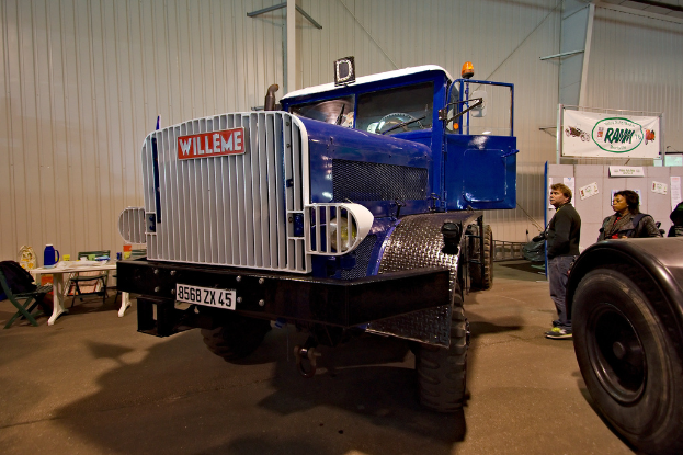 Ein Lastwagen steht in der Mitte mit ein paar Menschen, die rechts daneben stehen. Im Hintergrund ist eine Wand mit einem Tisch davor, auf dem ein Krug, eine Tasse und Deckel stehen, umgeben von Stühlen. Rechts daneben steht ein weiteres Fahrzeug mit einem sichtbaren Reifen.