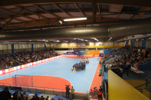 Eine große Halle voller Zuschauer bei einem Handballspiel, mit einem Netz auf der linken Seite, Anzeigetafeln im Hintergrund und Lampen und Rohren an der Decke.