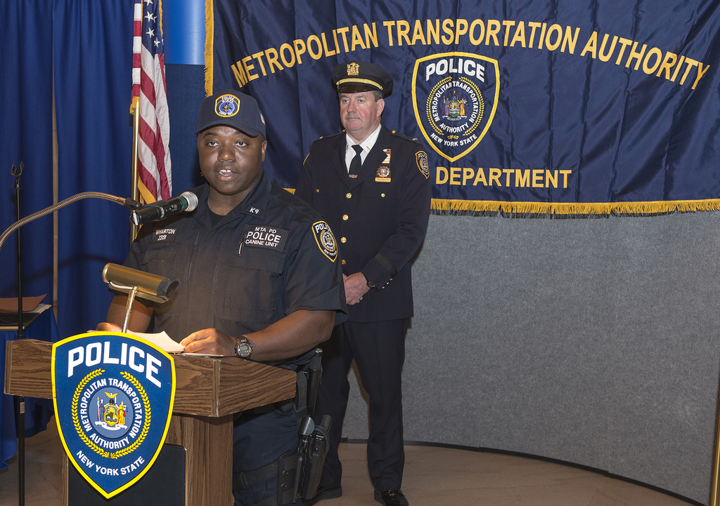 Zwei Polizisten stehen vor einem Rednerpult, einer hält Papers und spricht in ein Mikrofon, mit einer Fahne, einem Tisch und einer Tafel mit Symbolen und Text im Hintergrund.