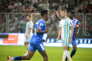 Ein Fussballspieler in einem blauen und weissen Trikot feiert mit erhobenen Armen, umgeben von einer Gruppe von Menschen in weissen und grünen Trikots, mit einem unscharfen Hintergrund, der ein weisses und blaues Brett und Geländer zeigt.