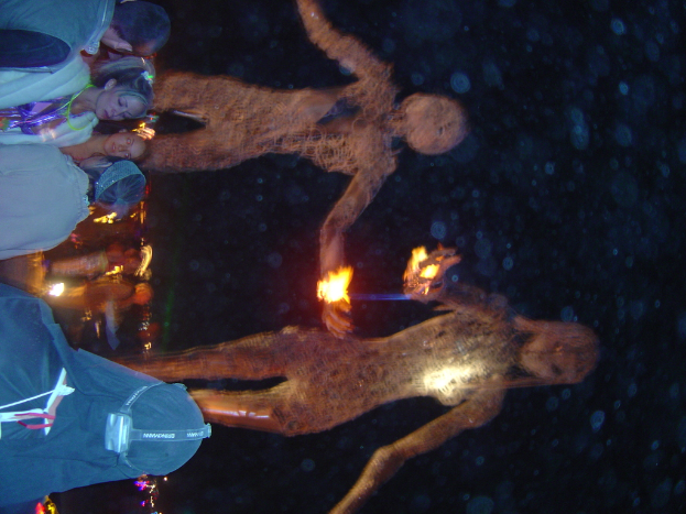 Mehrere Personen stehen auf der linken Seite des Bildes, dahinter brennende Statuen und Lichter im Hintergrund.