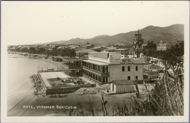 Schwarzes und weißes Foto eines Strandhotels, umgeben von Gebäuden, Bäumen, Wasser und Bergen mit klarem Himmel im Hintergrund.