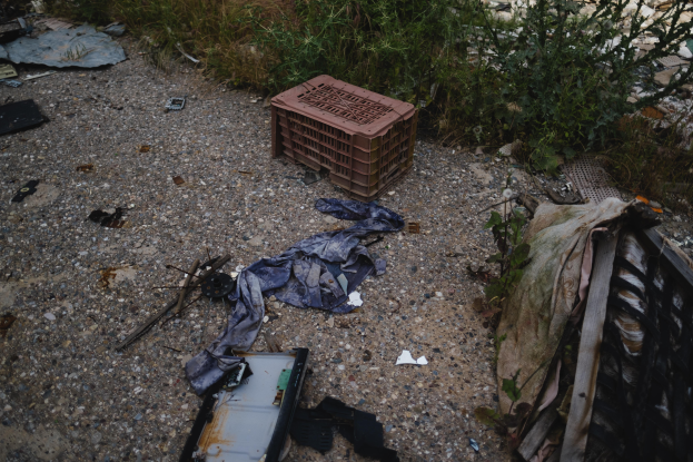 Ein Haufen Müll, einschließlich einer Plastikbox und Kleidung, liegt neben einem Zaun auf dem Boden einer Deponie, mit Pflanzen und Gras im Hintergrund.