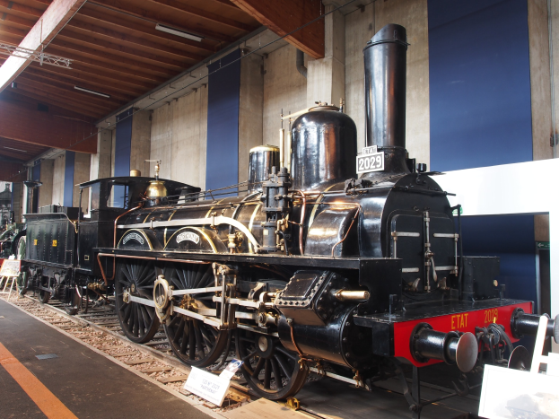Alte Dampflokomotive in einem Museum ausgestellt, umgeben von Eisenbahnschienen, Informationsplakaten, Ständen und anderen Ausstellungsstücken, mit einer Wand und beleuchteter Decke im Hintergrund.