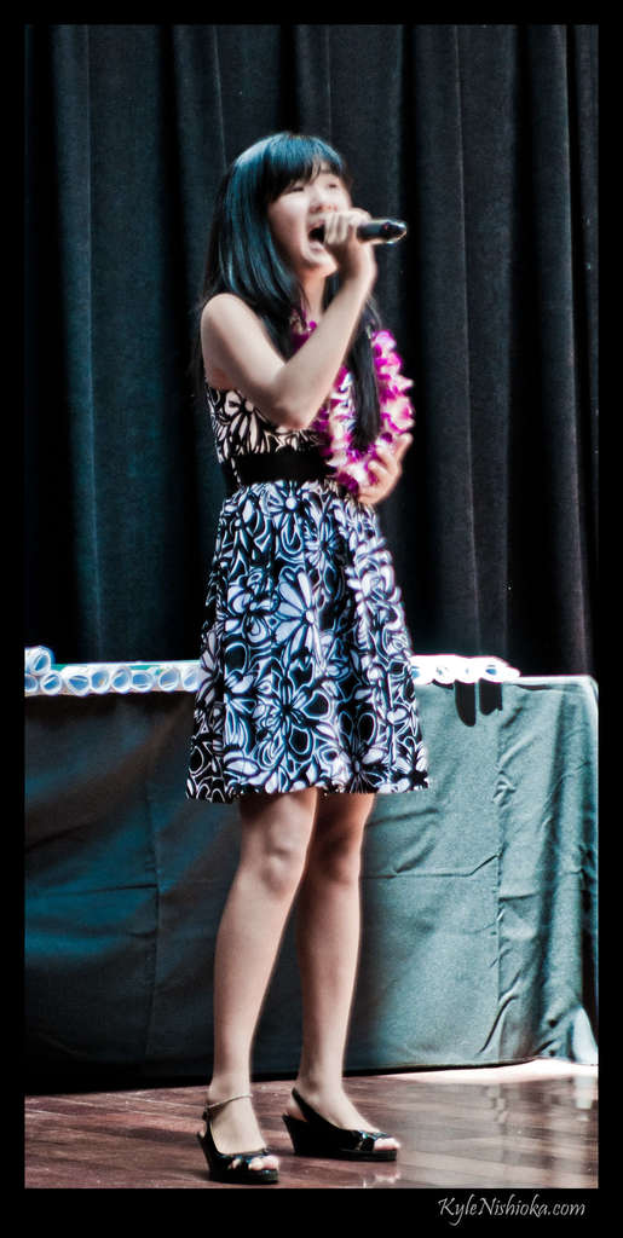 Eine Frau in einem bunten Kleid singt in ein Mikrofon auf der B├╝hne, mit einem Tisch mit Papieren und einem Vorhang im Hintergrund.