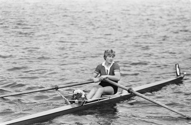 Eine Frau in einem weißen Hemd und blauen Shorts rudert ein Einerboot auf einem sonnigen Gewässer, wobei sie zwei Paddel verwendet und ihr Haar zu einem Pferdeschwanz gebunden hat.