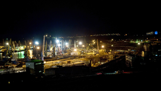 Eine nächtliche Stadtansicht mit beleuchteten Gebäuden, fahrenden Fahrzeugen auf einer Straße und Wasser im Vordergrund, vor einem dunklen Himmel.