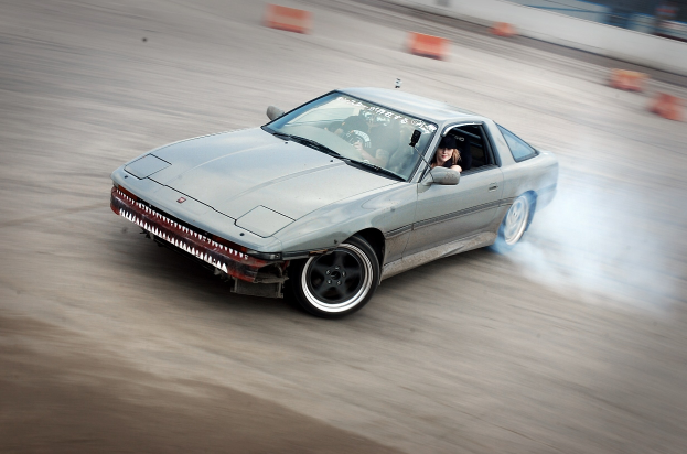 Eine Person in einem Helm fährt ein Auto auf einer Rennstrecke, umgeben von orangefarbenen Gegenständen, mit Rauch aus dem Fahrzeug gegen einen unscharfen Hintergrund.