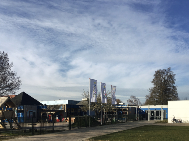 Großes Schulgebäude mit Metallzaun, Rasenfläche, Weg, Bäumen, Bannern, einem geparkten Fahrrad, nahen Häusern und einem bewölkten Himmel.
