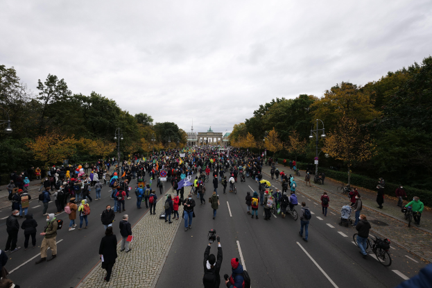 Eine große Gruppe von Menschen marschiert auf einer von Bäumen gesäumten Straße in Berlin, hält Kameras während einer Protestaktion, mit einem Gebäude und einem klaren Himmel im Hintergrund.