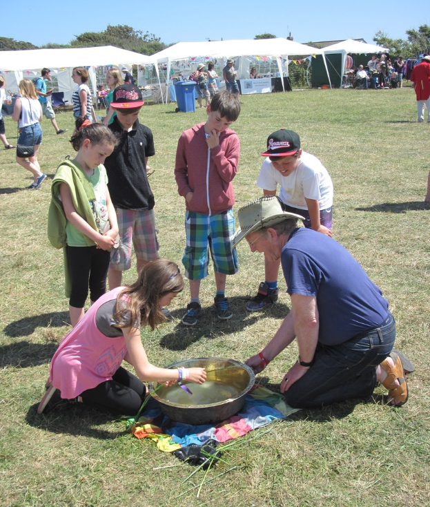 Zwei Personen knien auf dem Boden und arbeiten, mit Kindern im Vordergrund, Zelte, Menschen und ein Müllcontainer im Hintergrund unter einem Himmel.