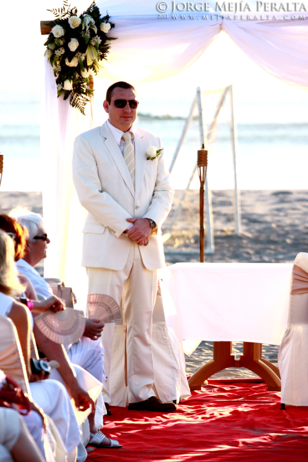 Ein Mann steht in der Mitte einer Zeremonie mit einem Strauß und einem Vorhang hinter ihm, während Menschen auf der linken Seite sitzen und ein Tisch mit einem Stuhl auf der rechten Seite ist, mit Pfählen, Sand und Wasser im Hintergrund unter einem sonnigen Himmel.