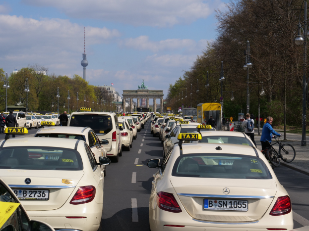 Eine Reihe von Taxis, die auf der Seite einer belebten Straße in Berlin, Deutschland, geparkt sind, mit Fahrzeugen, Radfahrern und Fußgängern, flankiert von Laternenmasten, Bäumen und Gebäuden, einschließlich eines Bogens und eines Turms, unter einem bewölkten Himmel.