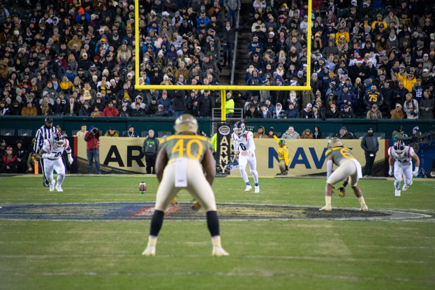 Ein Fußballspiel im Gange mit Spielern in Helmen und Sportbekleidung im Vordergrund und einer Zuschauermenge im Hintergrund, mit sichtbaren Pfosten und Texttafeln.