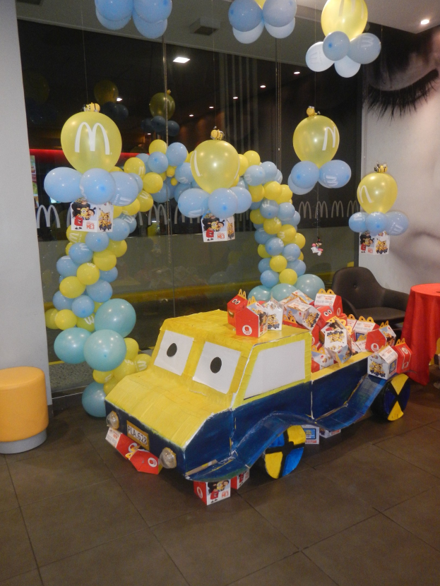 Eine McDonald's-Geburtstagsfeier mit einem gelben und blauen Lastwagen, der mit Ballons und anderen Dekoartikeln geschmückt ist, einem rot eingedeckten Tisch mit Gegenständen, Hockern und einer Glaswand im Hintergrund unter festlicher Deckenbeleuchtung.