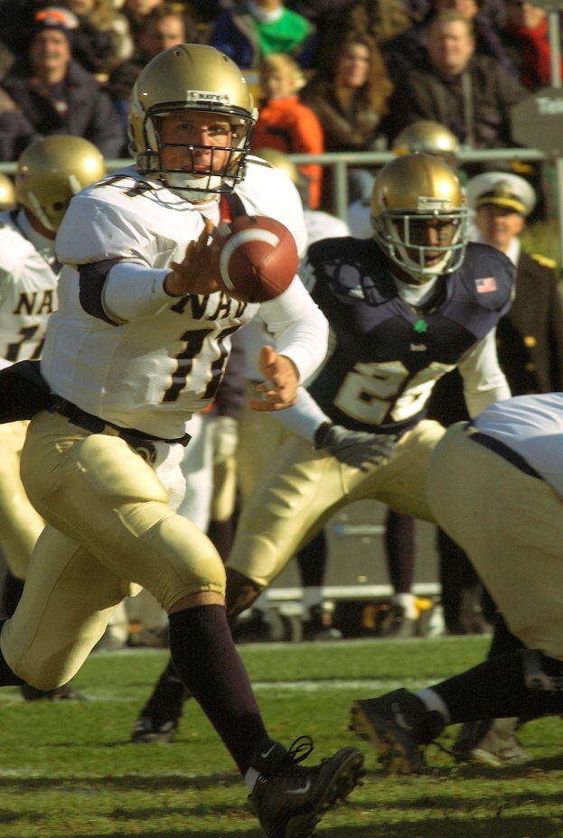 Ein Fussballspieler in einem Sportdress und Helm rennt mit dem Ball, während ihn andere Spieler verfolgen, mit einer Menge und einem Geländer im Hintergrund.