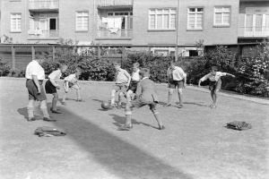 Schwarzes Bild von jungen Jungs, die auf einem Rasenfeld Fußball spielen, mit einem Ball in der Mitte, Gebäuden mit Fenstern, Geländern und Balkonen im Hintergrund und Pflanzen und Bäumen.