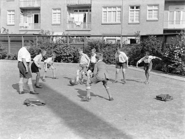 Schwarzes Bild von jungen Jungs, die auf einem Rasenfeld Fußball spielen, mit einem Ball in der Mitte, Gebäuden mit Fenstern, Geländern und Balkonen im Hintergrund und Pflanzen und Bäumen.