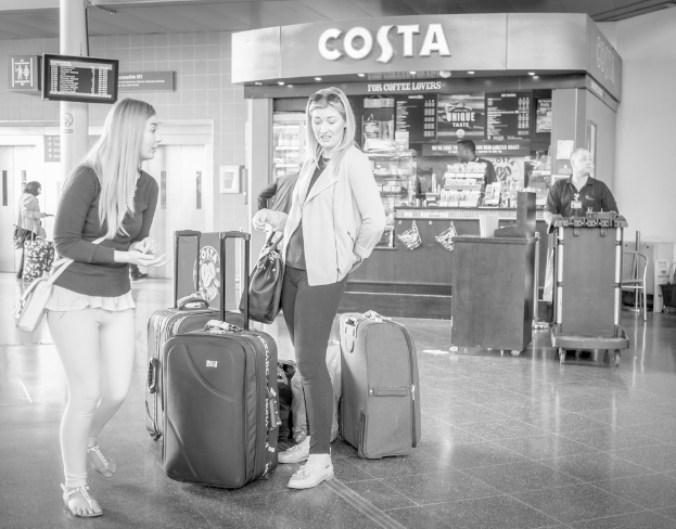Zwei Frauen mit Gepäcktaschen vor einem Café stehend, wahrscheinlich am Flughafen.