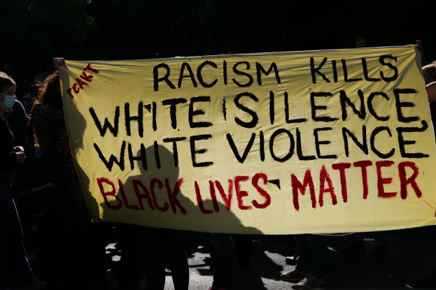 Eine Gruppe maskierter Menschen steht nachts auf einer Straße und hält ein Schild mit der Aufschrift "Rassismus tötet Weiße Stille, weiße Gewalt, Schwarze Leben zählen".