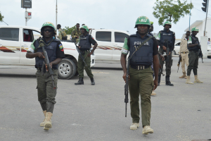 Eine Gruppe von Polizisten in Helmen und mit Waffen in der Hand geht eine Straße entlang mit Fahrzeugen, Laternenmasten, Verkehrsampeln, Texttafeln, Bäumen, Gebäuden und einem bewölkten Himmel im Hintergrund.