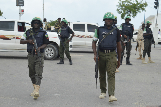 Eine Gruppe von Polizisten in Helmen und mit Waffen in der Hand geht eine Straße entlang mit Fahrzeugen, Laternenmasten, Verkehrsampeln, Texttafeln, Bäumen, Gebäuden und einem bewölkten Himmel im Hintergrund.