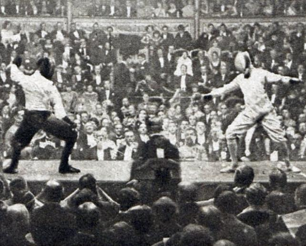 Zwei Männer fechten in einem Stadion, einer mit einem Schwert und der andere mit einem Schild, vor einem jubelnden Publikum im Hintergrund, in Schwarz-Weiß abgebildet.