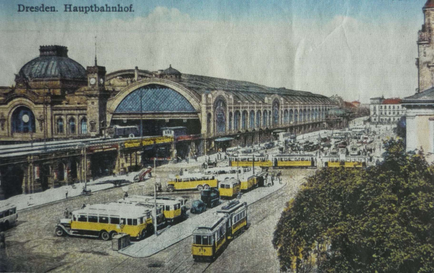 Ein altes Postkartenmotiv zeigt einen belebten Dresdner Bahnhof mit Fahrzeugen, Menschen, Bäumen, Gebäuden und einem bewölkten Himmel, mit der Aufschrift "Dresden Hauptbahnhof" oben.
