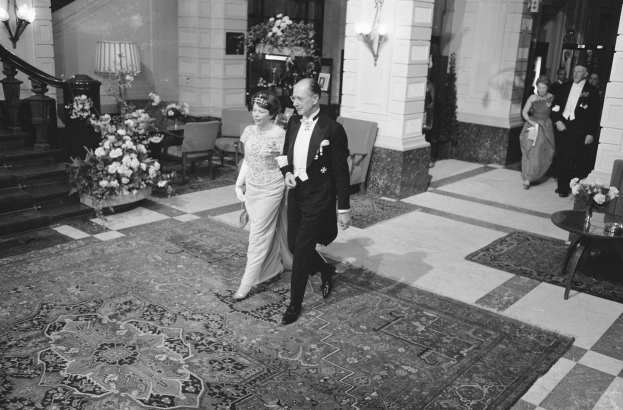 Ein Schwarz-Weiß-Bild einer Braut und eines Bräutigams, die durch die Hotellobby gehen, umgeben von Menschen, Möbeln und Dekoration.