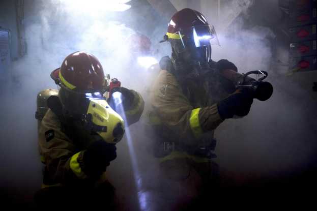 Zwei Feuerwehrleute in Schutzanzügen spritzen Wasser auf eine rauchgefüllte Wand, während links ein Schild mit Text zu sehen ist.