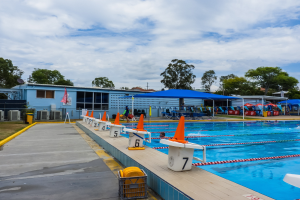 Großer Außenpool mit schwimmenden Menschen, umgeben von Bahnmarkierungen, Verkehrskegeln, Stühlen, Sonnenschirmen, einem Gebäude mit Fenstern, einer Flagge, Bäumen und einem bewölkten Himmel.