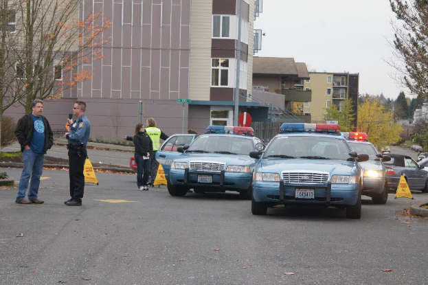 Autos auf einer Straße mit vier Personen in der Nähe, Gebäude mit Fenstern im Hintergrund, Bäume und Notfallwarndreiecke.