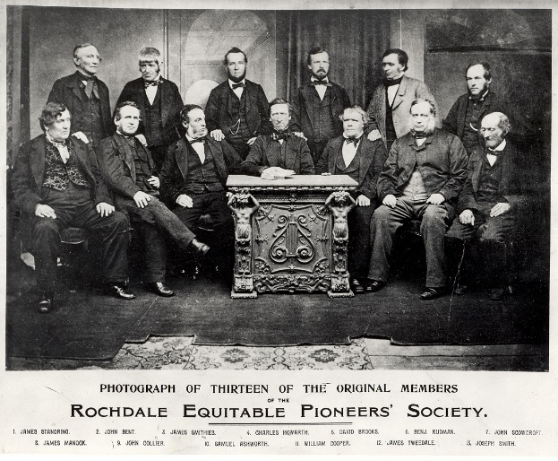 Eine Gruppe von Männern in Blazern und Krawatten, die um einen Tisch sitzen, mit der Aufschrift "Rochdale Equitable Pioneers Society" unten.