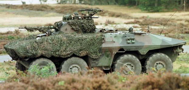 Militärischer Mannschaftstransporter mit einer montierten großen Waffe, der auf einer Straße neben einem Wald fährt.