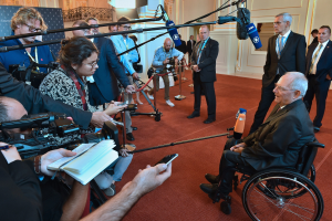 Ein Mann im Rollstuhl in Anzug und Krawatte von Reportern umgeben, die Kameras und Mikrofone halten, mit einem Banner und einer Wand im Hintergrund.