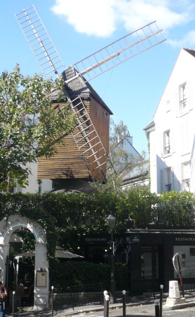 Eine Windmühle, mehrere Gebäude, Bäume, Metallstangen und ein Zaun in einer ländlichen Umgebung.