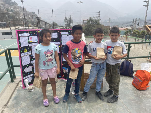 Eine Gruppe von Kindern, die auf einem Basketballfeld stehen und Papiertüten halten, mit Tüten und Gegenständen rechts und einer Tafel mit Text links, vor dem Hintergrund von Zäunen, Pfosten, Straßenlaternen, Bäumen, Gebäuden, Hügeln und dem Himmel.