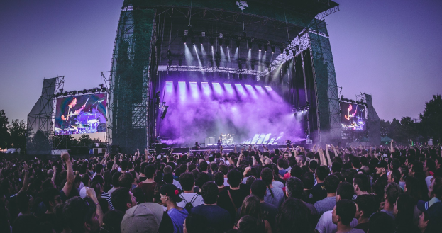 Eine große Menschenmenge schaut bei einem Musikfestival auf der Bühne mit Instrumenten und Bildschirmen, die vor einem Gebäude, Bäumen und einem klaren blauen Himmel aufgebaut sind.