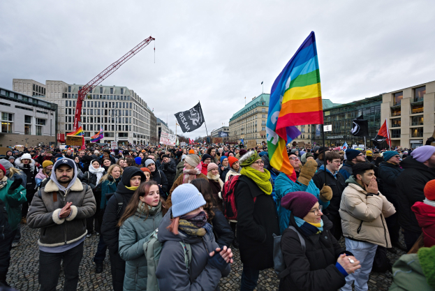 Eine große Gruppe von Menschen mit Fahnen und Schildern, darunter "Lgbtq+ Demonstrationszug in Berlin", vor einem Gebäude mit anderen Strukturen und einem bewölkten Himmel im Hintergrund.