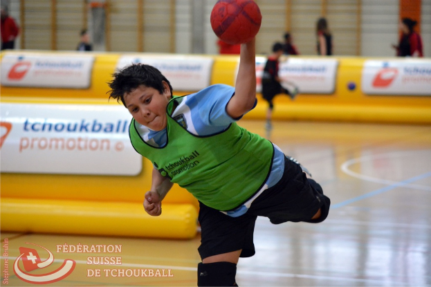 Ein Junge in weißem Hemd und dunkler Hose wirft einen Ball mit seiner linken Hand, mit einem gelben Luftballon und mehreren Menschen im Hintergrund.