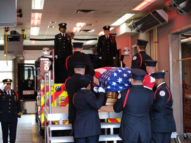 Eine Gruppe uniformierter Männer trägt einen Sarg mit einer amerikanischen Flagge, umgeben von ein paar Leuten mit Mützen und einem Feuerwehrauto sowie Deckenleuchten im Hintergrund.