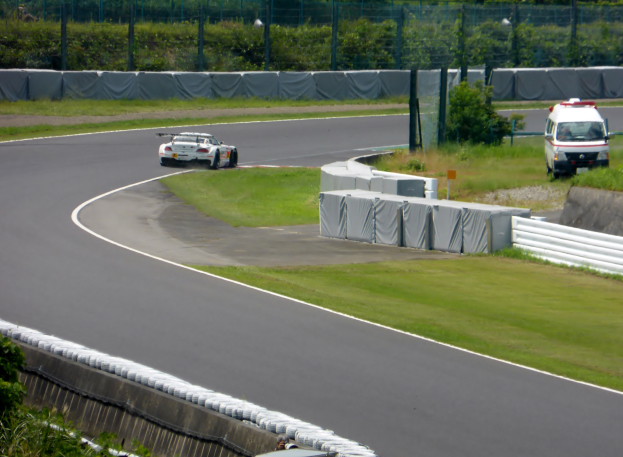 Ein Rennwagen fährt eine Rennstrecke entlang, im Hintergrund ein Polizeiwagen, umgeben von Grünflächen und Absperrungen, mit Text am unteren Bildrand.