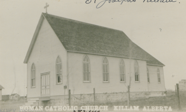 Ein Schwarz-Weiß-Foto der St. Joseph's Lutheran Church in Killam, Alberta, das ein Haus mit einem Dach, Fenstern, Türen und einem Kreuz auf dem Dach zeigt, das von einem Zaun, Gras und anderen Häusern im Hintergrund umgeben ist, mit Text oben und unten auf dem Bild.