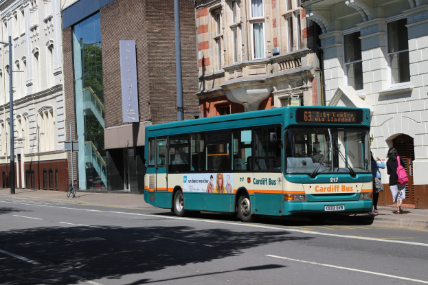 Blauer Bus fährt auf einer Straße mit hohen Gebäuden, zwei Fußgängern auf dem Gehweg und einem Fahrrad im Hintergrund.