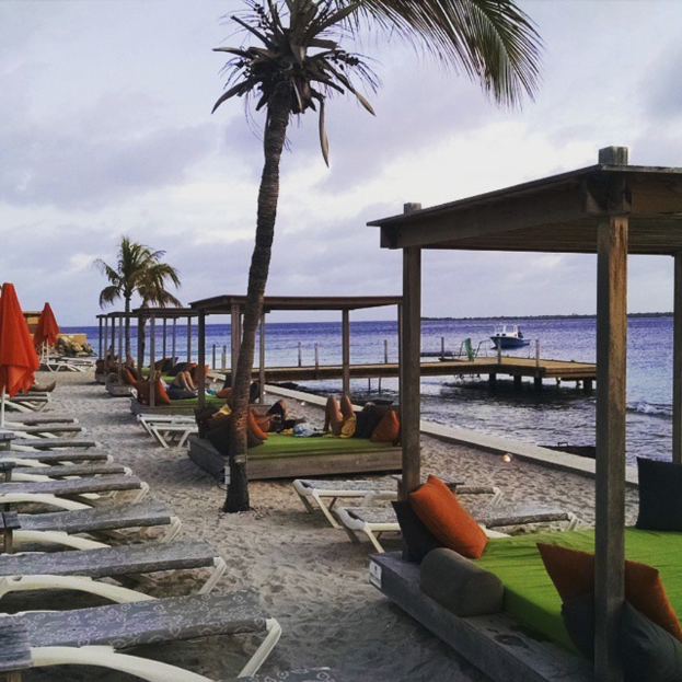 Eine Meeresansicht mit Stühlen, Betten, Bäumen, Sonnenschirmen auf dem Sand, einem Boot auf dem Wasser im Hintergrund und einer Gruppe von Menschen, die auf einem der Betten sitzen, unter einem bewölkten Himmel.