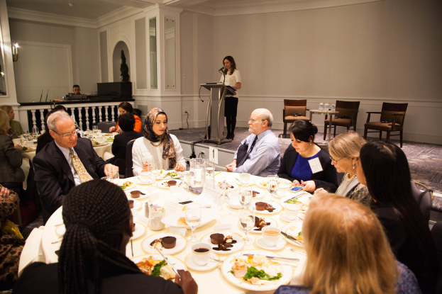 Eine Gruppe von Menschen sitzt um einen Esstisch mit Tellern, Besteck und Trinkgefäßen, während eine Frau in der Nähe eines Podiums mit einem Mikrofon im Hintergrund steht.