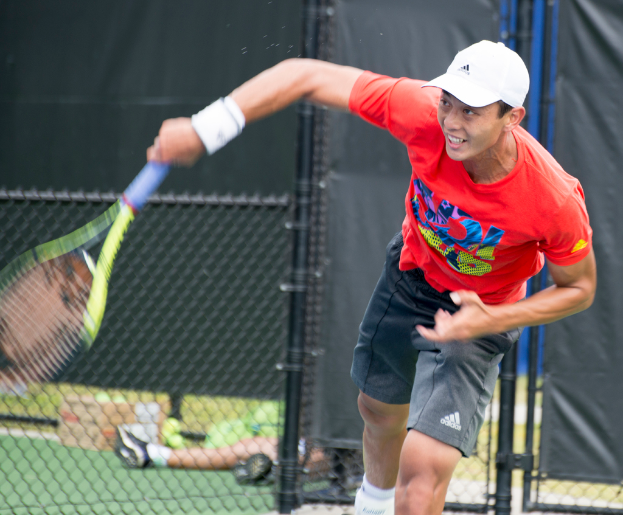 Ein Mann in roter Jacke und weißer Kappe spielt Tennis, hält einen Schläger und hat einen Maschendrahtzaun hinter sich; im Hintergrund sitzt eine Person auf dem Boden neben einer Box und Tennisbällen.