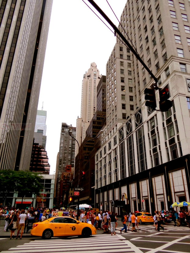 Eine Stadtansicht mit Gebäuden, Ampeln, Fenstern, wenigen Passanten, Autos und Himmel.