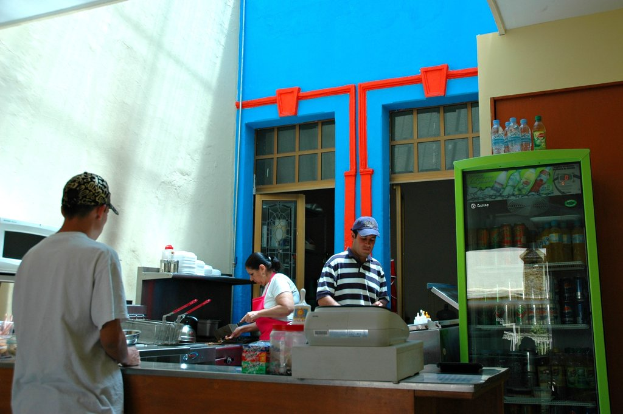 Drei Personen arbeiten in einer Küche, wobei ein Kühlschrank auf der rechten Seite des Bildes zu sehen ist.