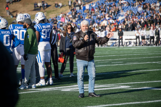 Ein Mann in einer schwarzen Jacke, blauen Jeans und schwarzen Schuhen steht auf einem Fußballfeld und hält ein Mikrofon, mit einer Menge Menschen im Hintergrund, einige tragen Helme.